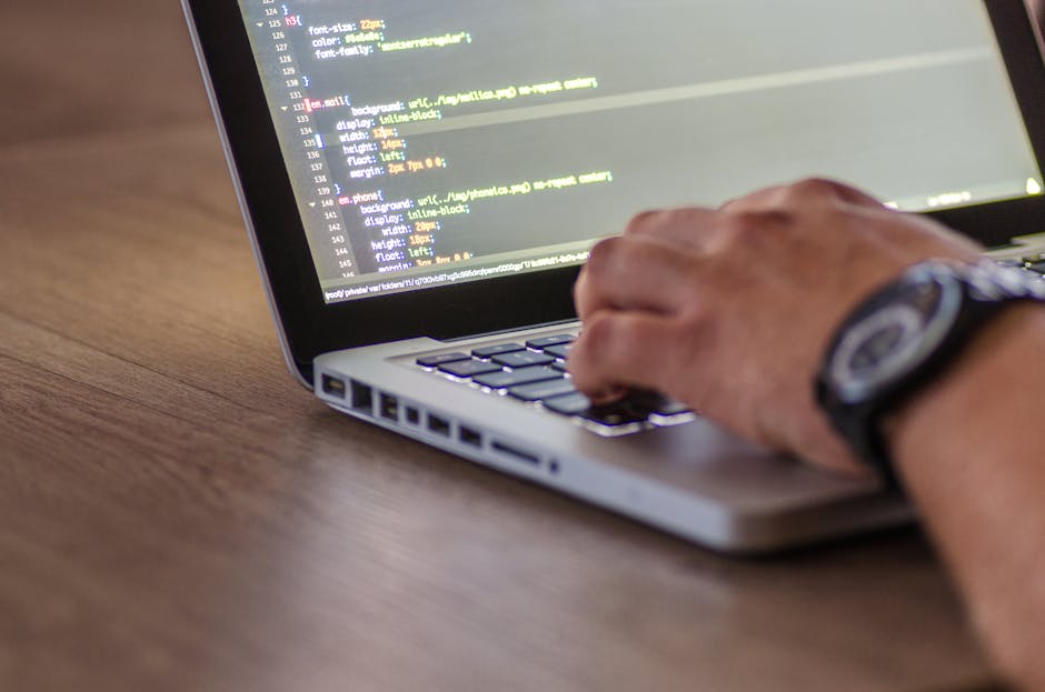 Close-up of a person's hand typing on a laptop keyboard placed on a wooden desk. The laptop screen displays lines of programming code with syntax highlighted in various colors. The person is wearing a