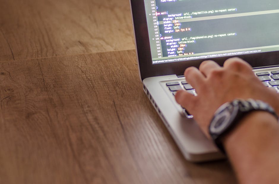 Close-up of a person's hand typing on a laptop keyboard placed on a wooden desk. The laptop screen displays lines of programming code with syntax highlighted in various colors. The person is wearing a