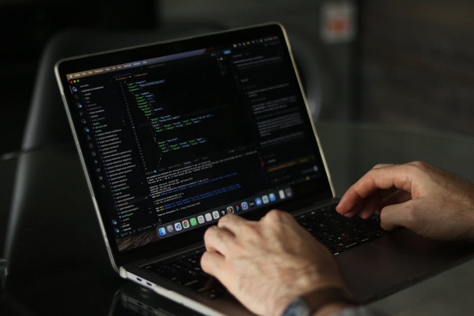 A person working on a laptop with a black screen displaying lines of colorful code in green, blue, and white, situated on a dark table. The individual's hands are visible, with the right hand poised o